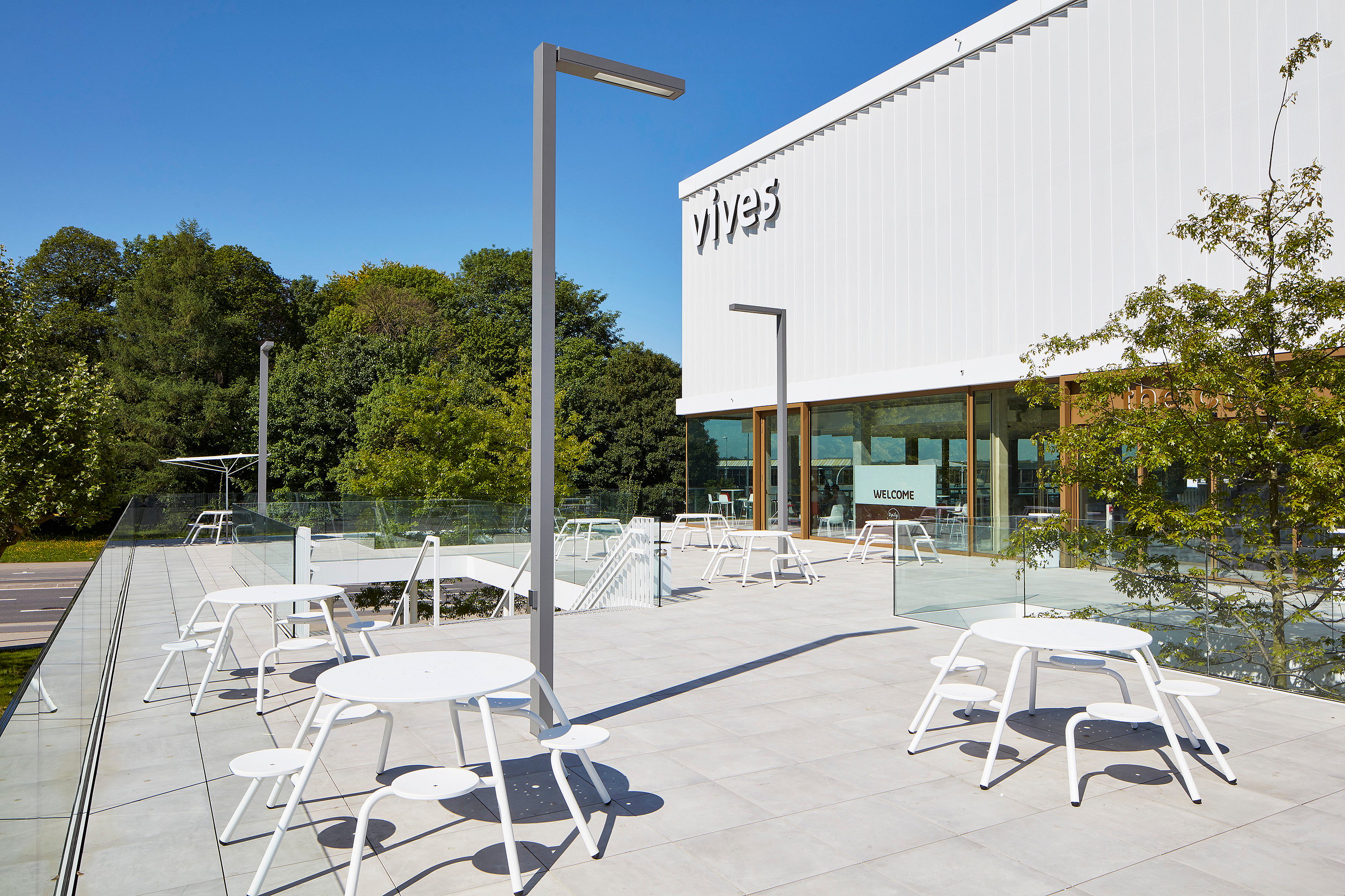 Wide sunlit terrace at the Vives campus with several white round picnic tables and stools, glass railings, tall light poles, and the modern white campus building in the background.