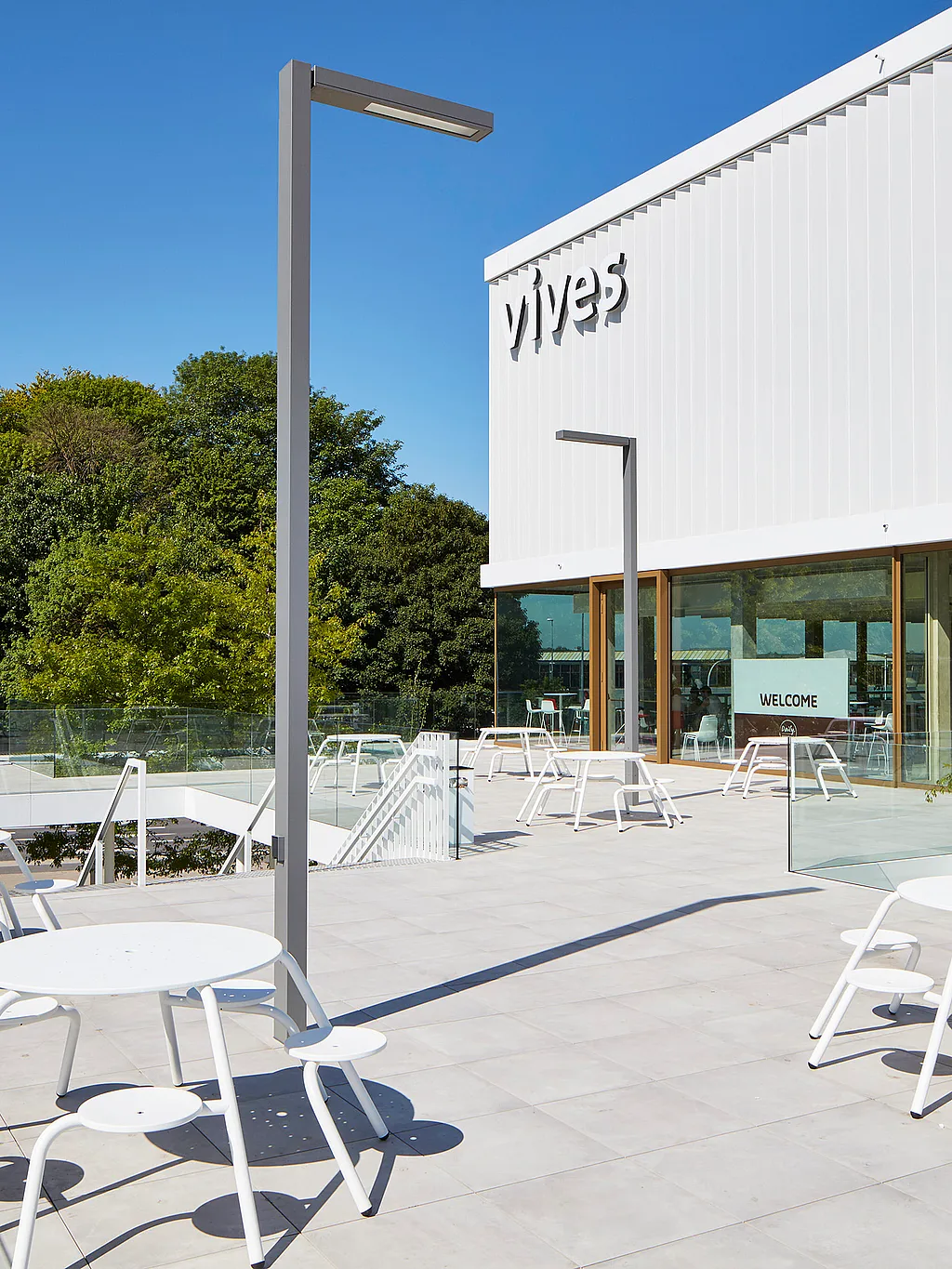 Wide sunlit terrace at the Vives campus with several white round picnic tables and stools, glass railings, tall light poles, and the modern white campus building in the background.
