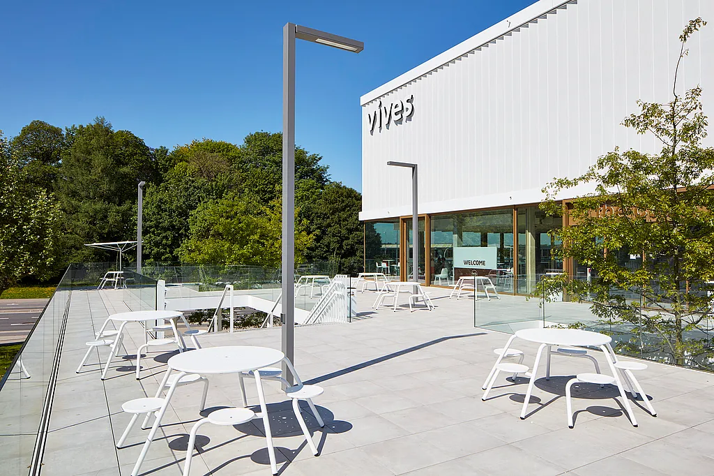 Wide sunlit terrace at the Vives campus with several white round picnic tables and stools, glass railings, tall light poles, and the modern white campus building in the background.