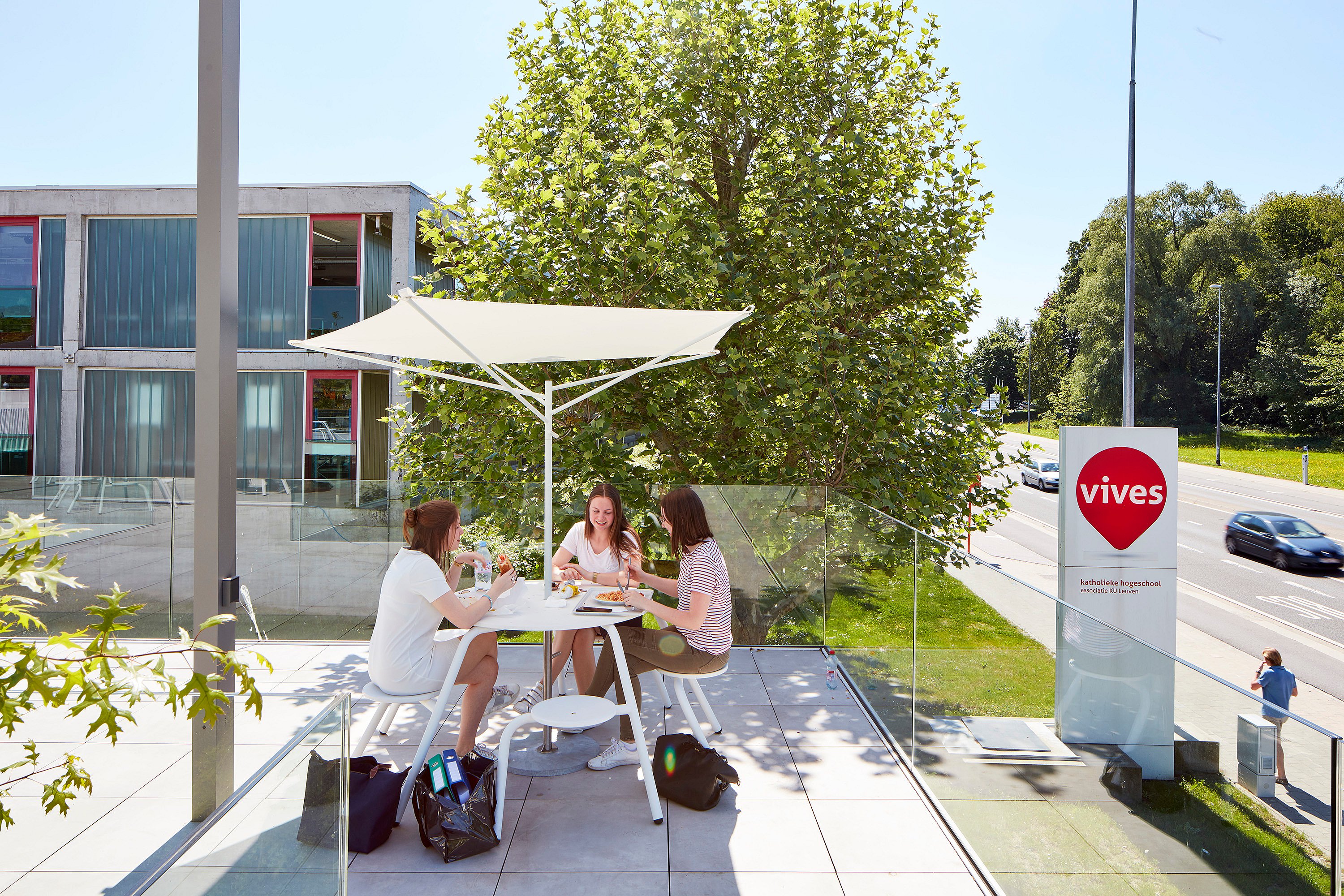   Drei Personen sitzen an einem weißen Tisch auf der Terrasse neben einem großen Baum. Rechts sind das Schild des Vives-Campus und die Straße zu sehen.