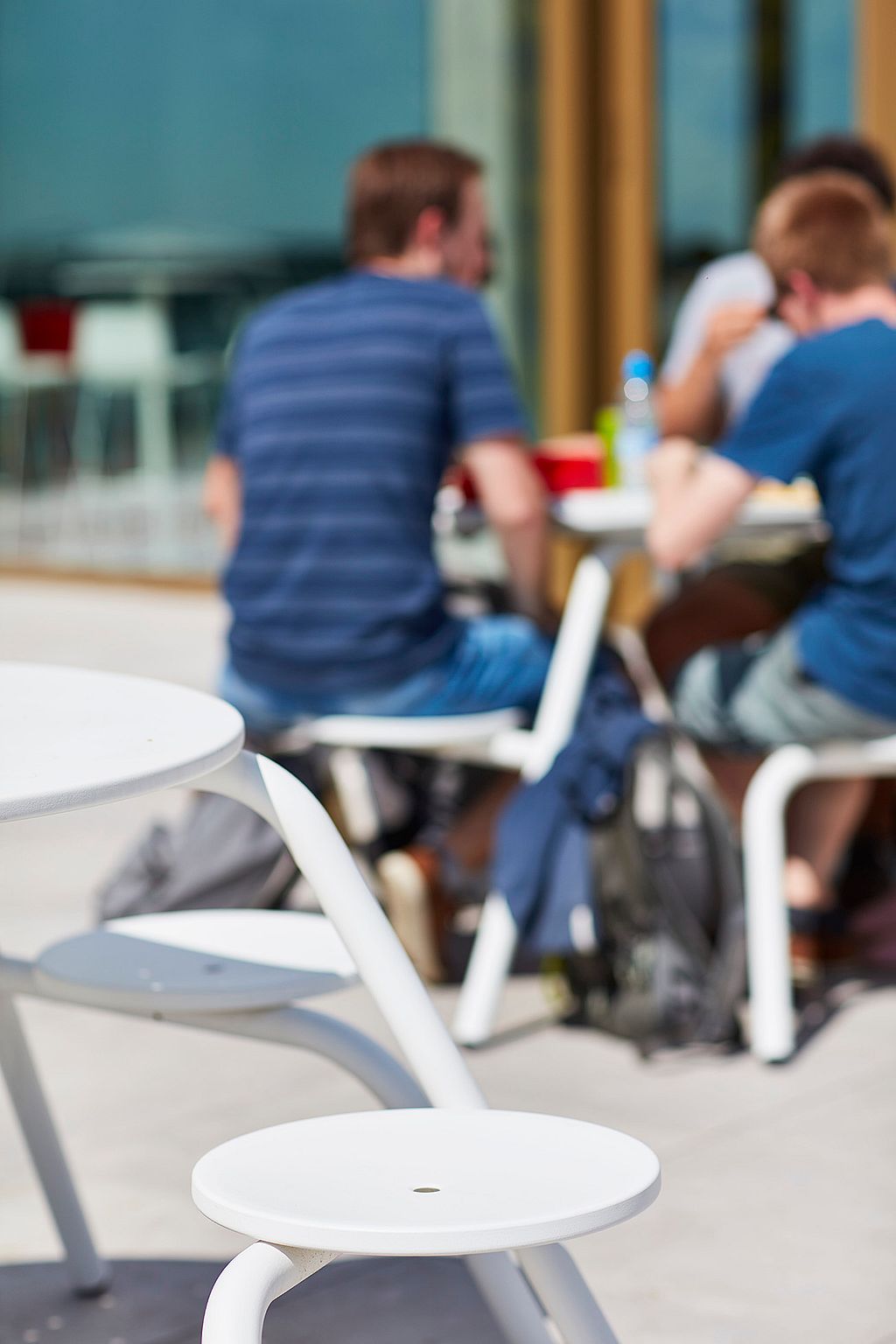 Studierende haben sich an mehreren weißen Picknicktischen auf einer geräumigen Terrasse vor dem Campus-Eingang versammelt und genießen die Sonne beim Essen und Plaudern.