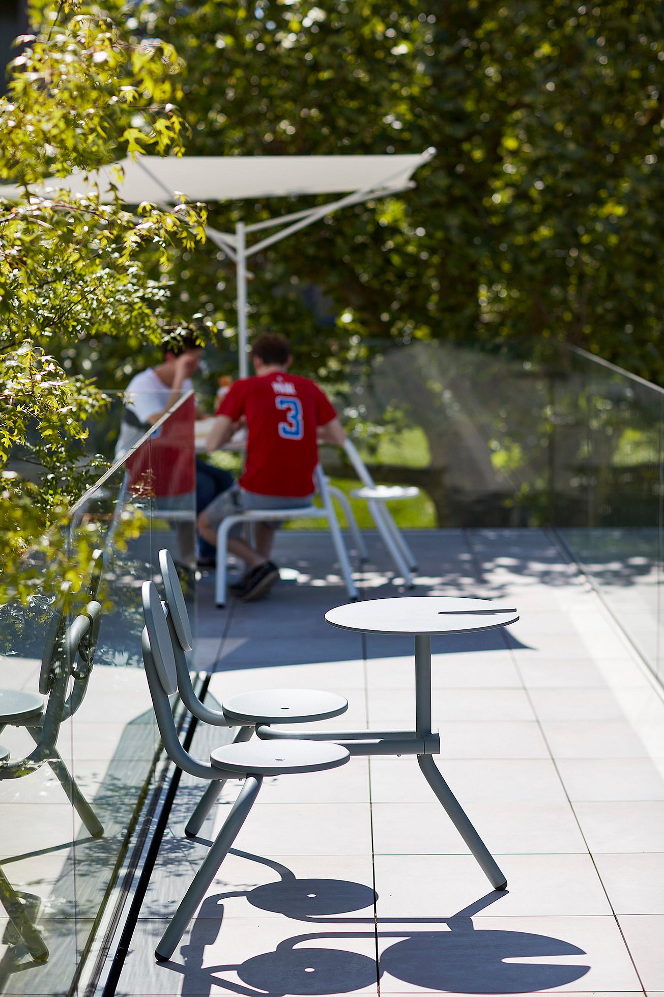   Nahaufnahme eines weißen Tisches mit daran befestigten runden Hockern neben einer Glasbalustrade. Im verschwommenen Hintergrund sitzen zwei Personen unter einem weißen Sonnensegel.
