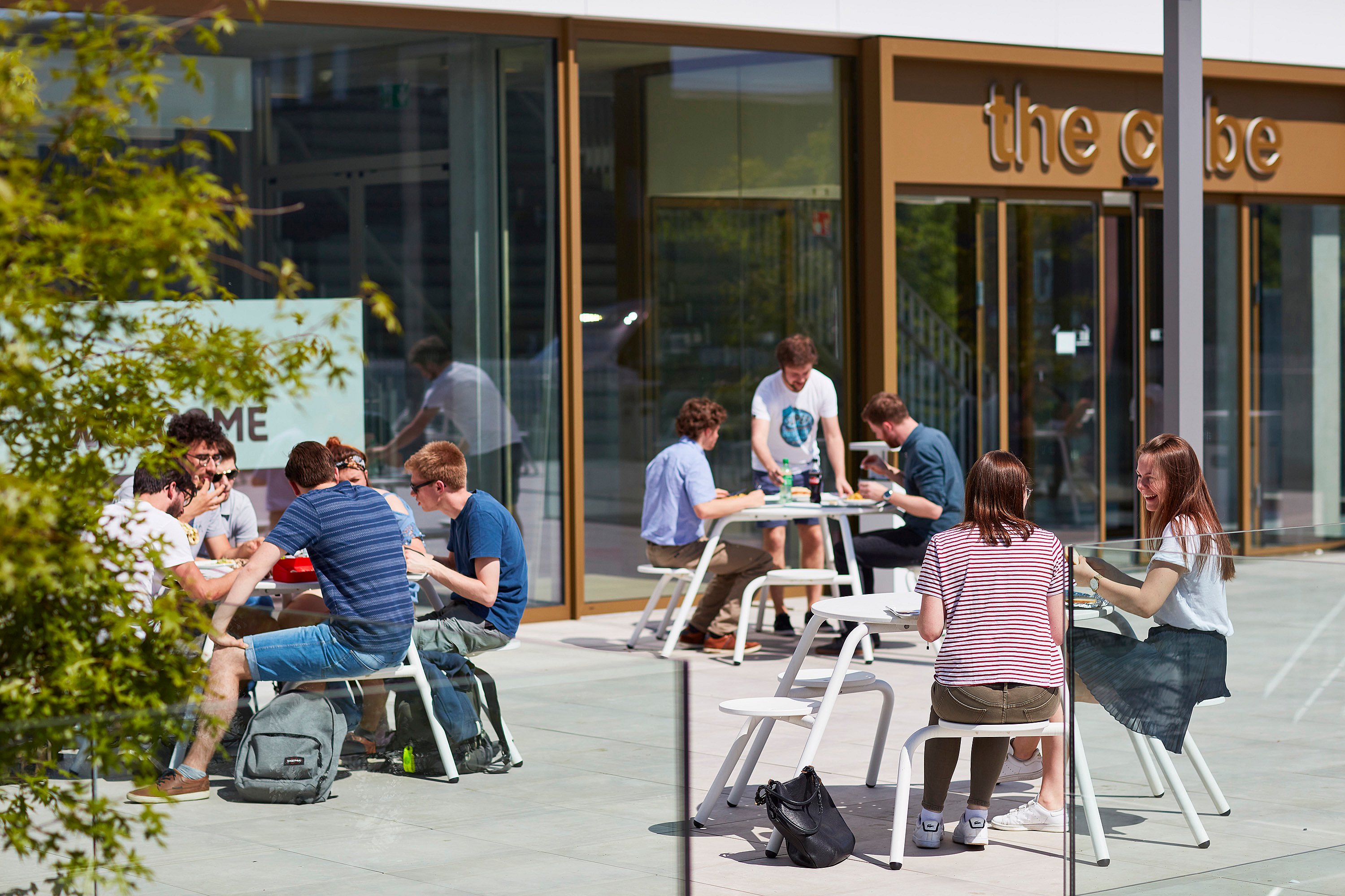   Studierende haben sich an mehreren weißen Picknicktischen auf einer geräumigen Terrasse vor dem Campus-Eingang versammelt und genießen die Sonne beim Essen und Plaudern.
