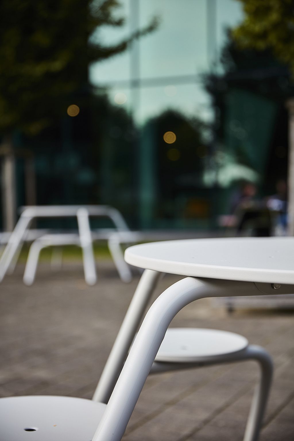 Detailweergave van een witte Virus-picknicktafel met bijbehorende krukjes op het geplaveide campusterras, met het gebouw en andere zitmeubelen vaag wazig op de achtergrond.