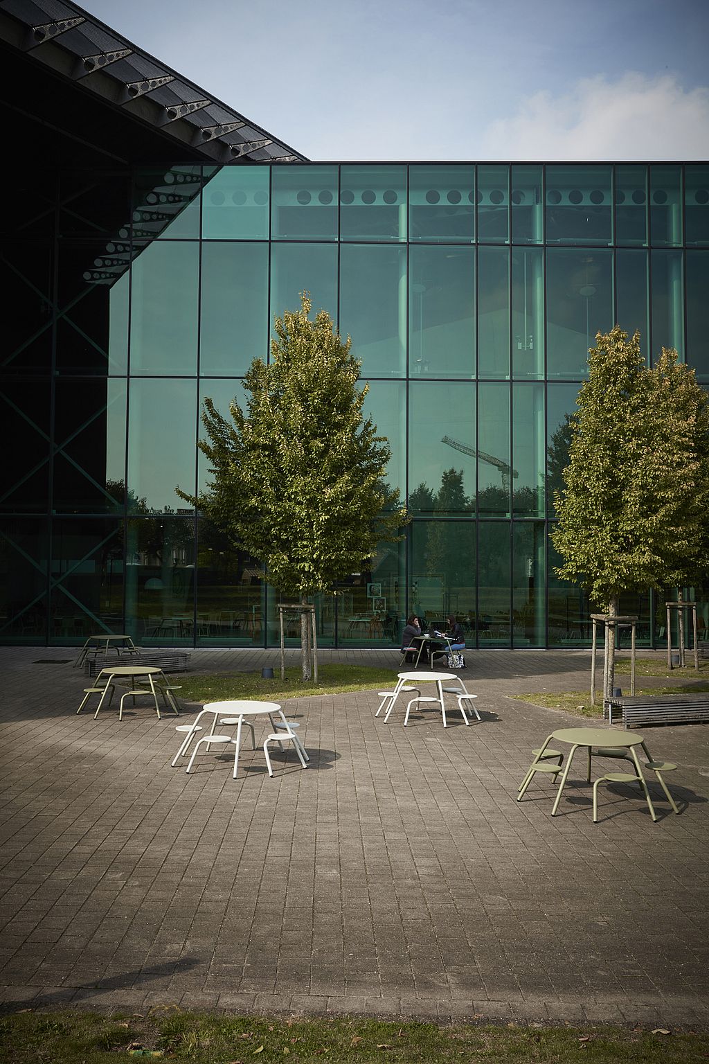 Breed overzicht van een universiteitsgebouw met een glazen gevel en ronde witte en olijfgroene Virus-picknicktafels verspreid over de open binnenplaats onder hoge bomen.