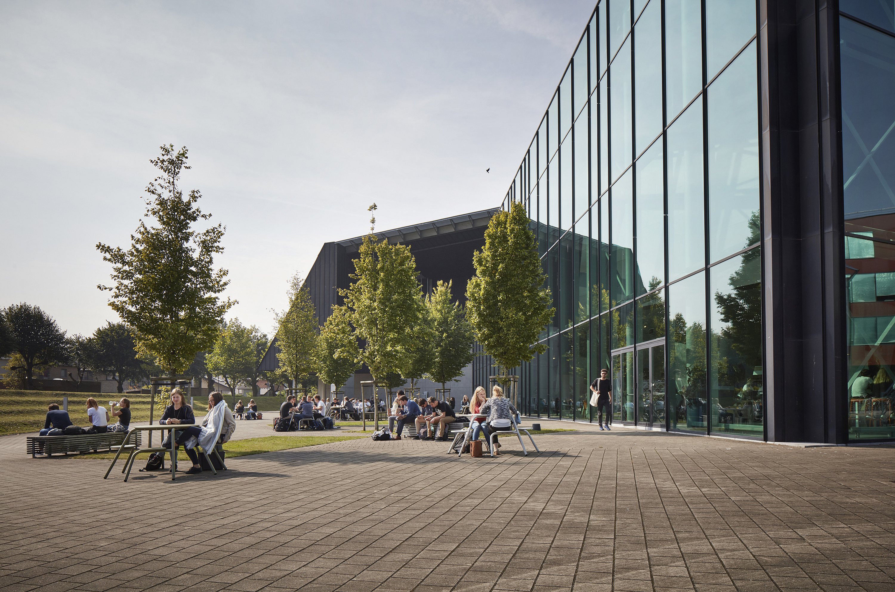   Studenten zitten te praten rond ronde picknicktafels op een breed geplaveid terras naast een modern glazen campusgebouw op een zonnige dag.