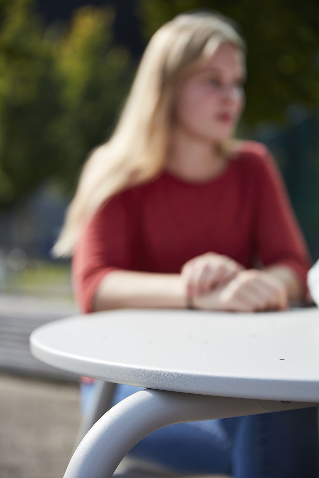 Close-up van een witte ronde picknicktafel met gebogen poten, scherp in beeld, met een zittend persoon wazig op de achtergrond.