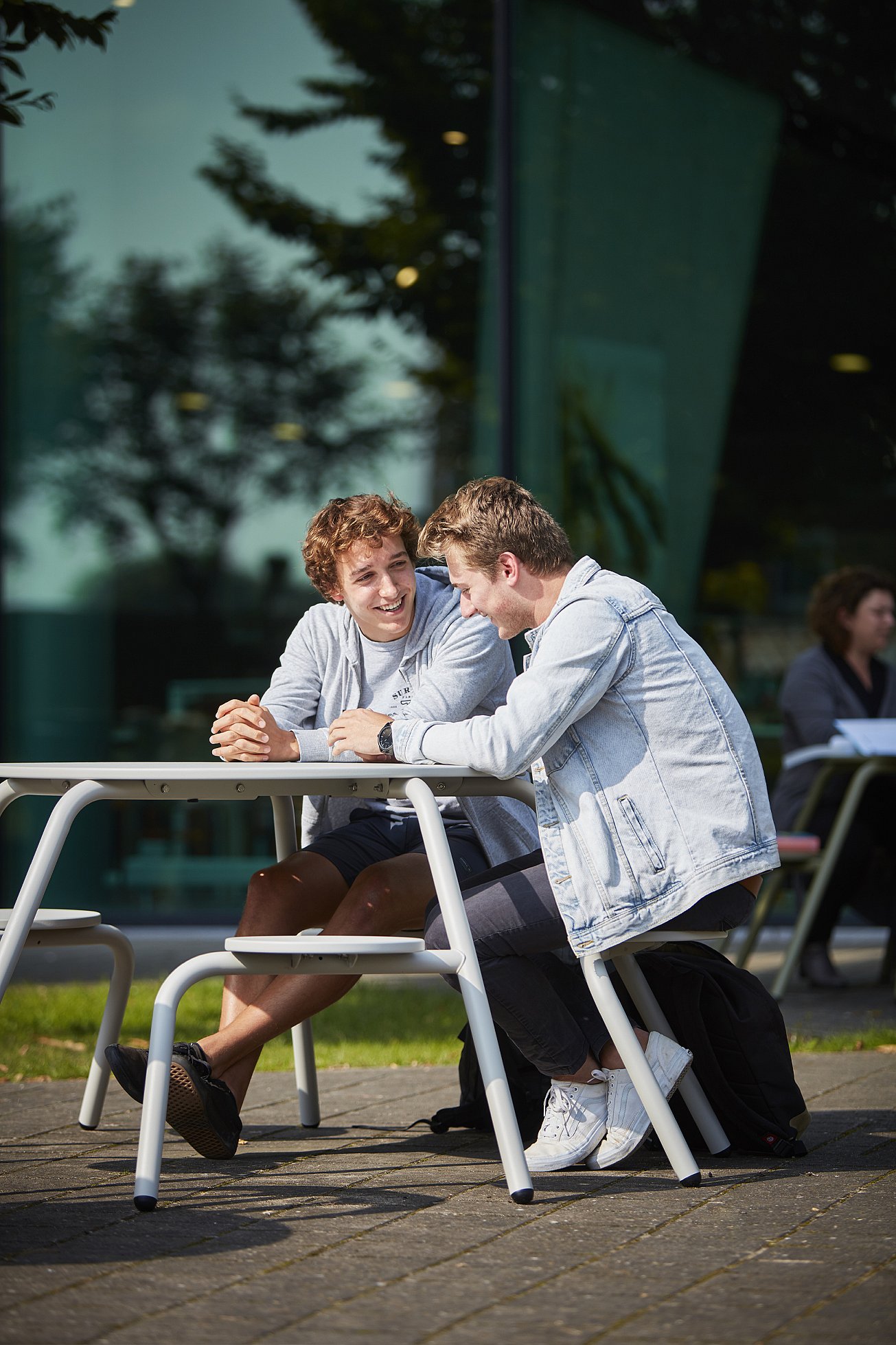   Twee studenten zitten dicht bij elkaar aan een witte picknicktafel voor een glazen wand, pratend en lachend in de zon.