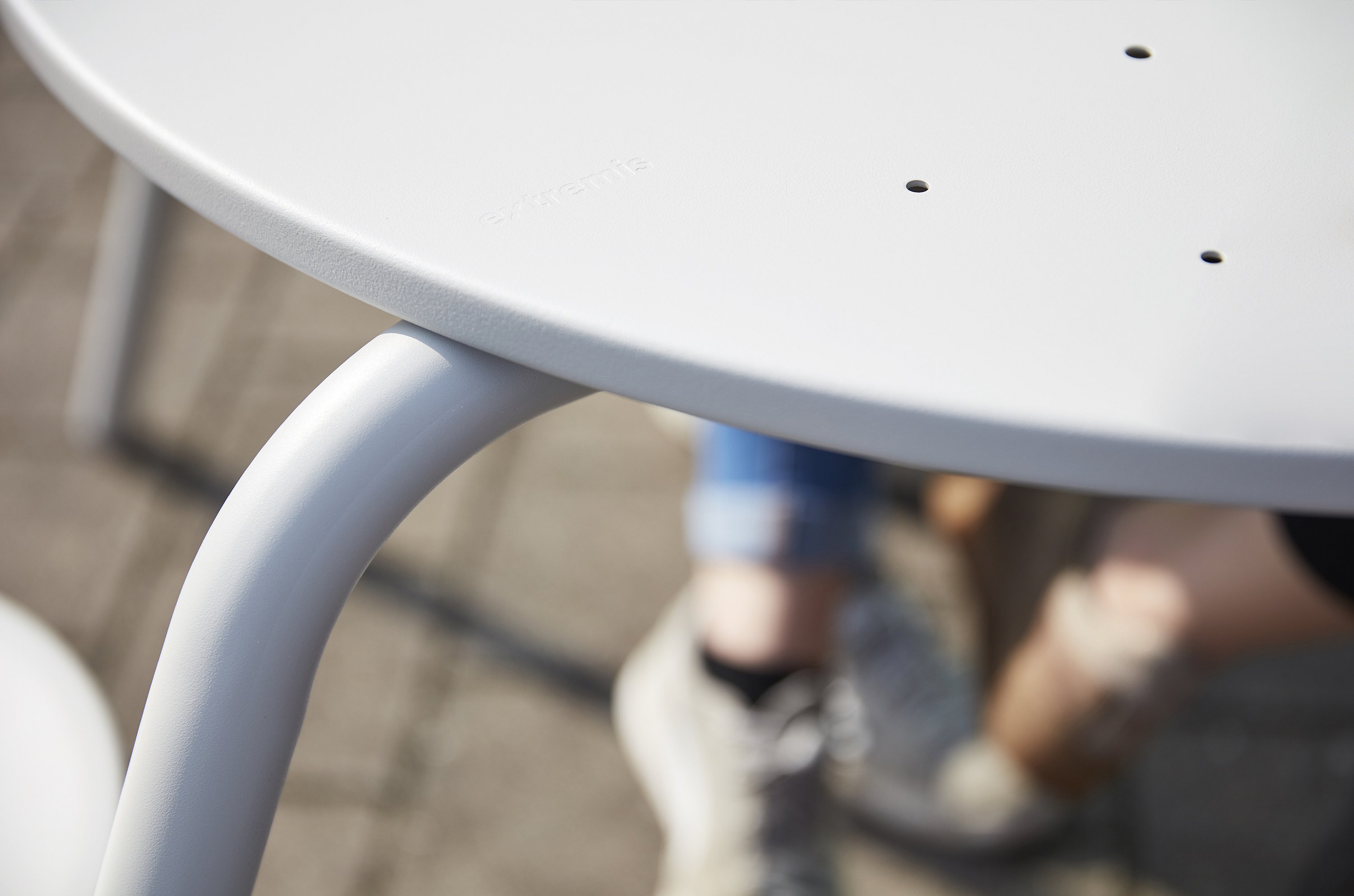   Close-up van de witte tafelrand en gebogen poot van een Virus-picknicktafel, met de benen en schoenen van een persoon wazig op de achtergrond.