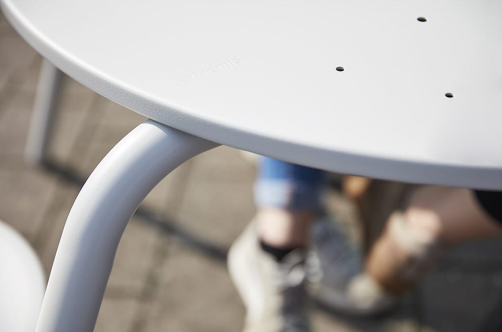 Close-up van de witte tafelrand en gebogen poot van een Virus-picknicktafel, met de benen en schoenen van een persoon wazig op de achtergrond.