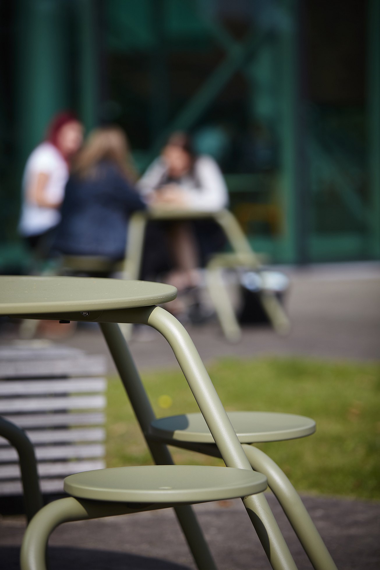   Close-up van een rietgroene Virus-picknicktafel met ronde krukjes, met een kleine groep mensen die aan een andere tafel zit, onscherp op de achtergrond.