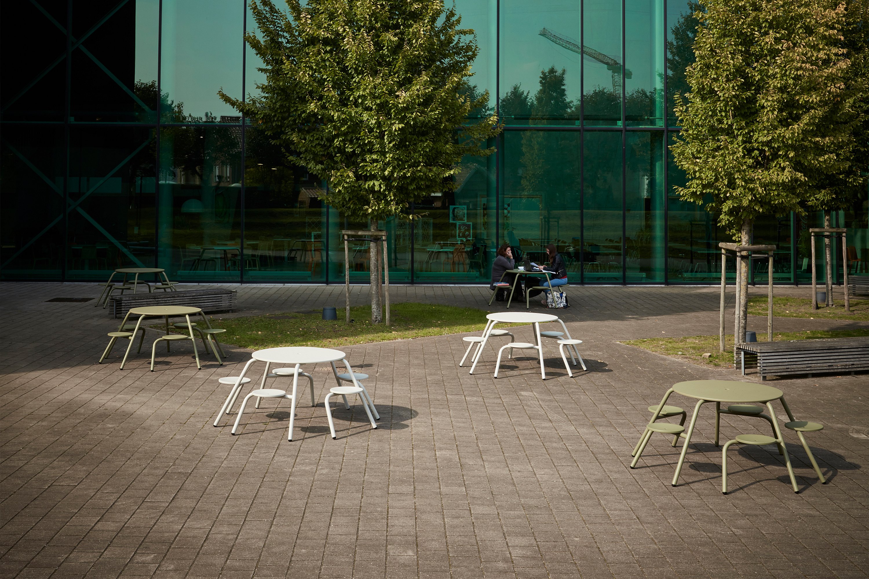 Plusieurs tables de pique-nique rondes Virus, blanches et vert olive, sont disposées dans une cour pavée du campus, avec de jeunes arbres et une haute façade vitrée de l'université en arrière-plan.