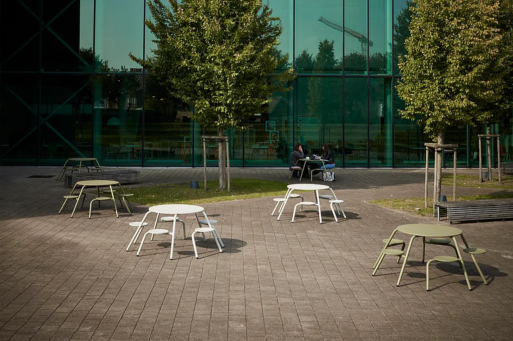 Plusieurs tables de pique-nique rondes Virus, blanches et vert olive, sont disposées dans une cour pavée du campus, avec de jeunes arbres et une haute façade vitrée de l'université en arrière-plan.