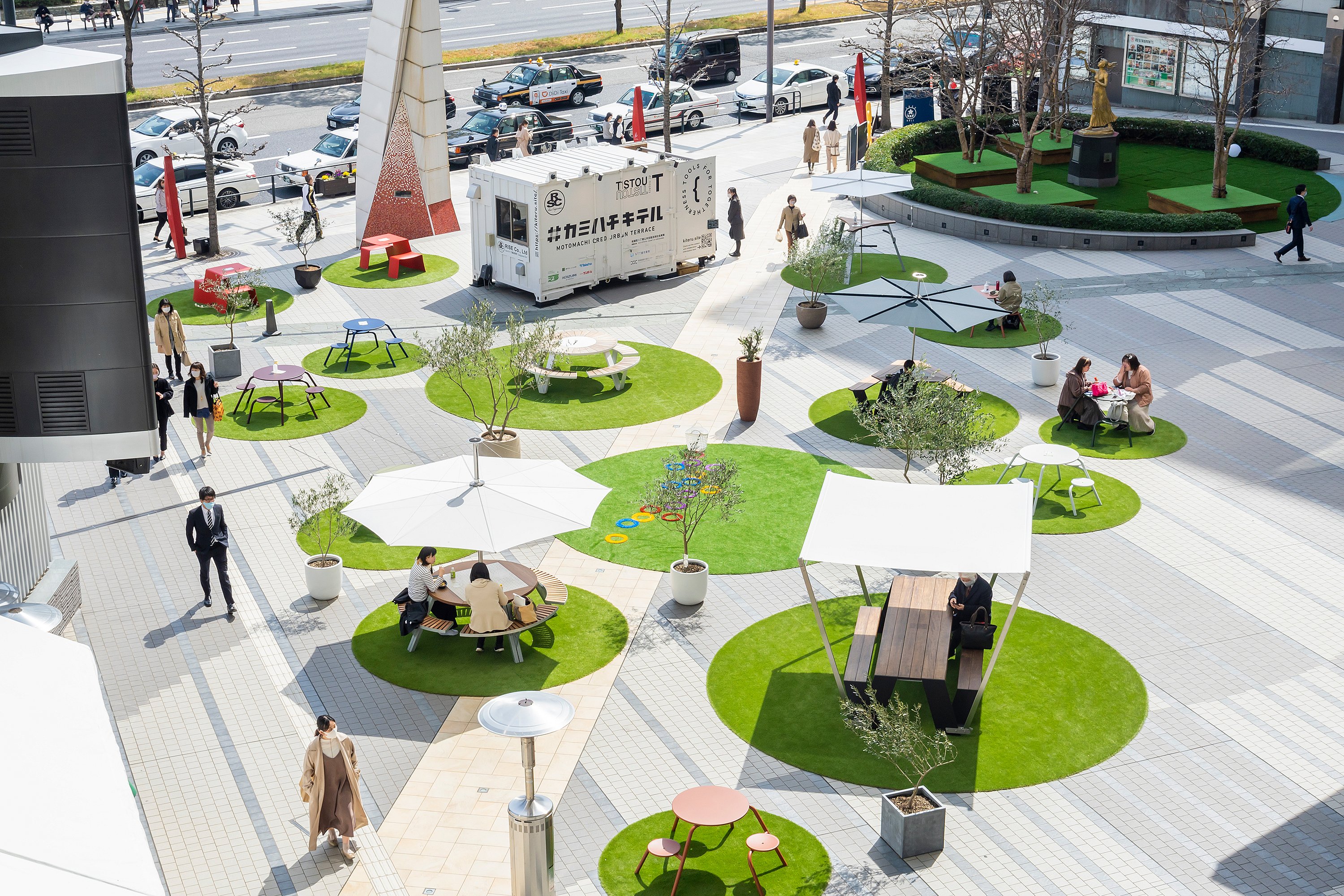 Bovenaanzicht van een openbaar plein in Hiroshima met heldere ronde stukken kunstgras, elk ingericht met verschillende buitentafels, banken, parasols en schaduwstructuren om op afstand geplaatste zitjes voor bezoekers te creëren.