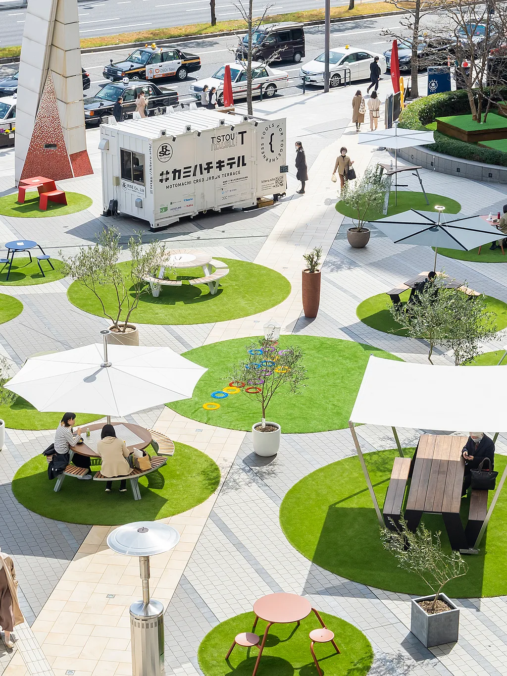 Bovenaanzicht van een openbaar plein in Hiroshima met heldere ronde stukken kunstgras, elk ingericht met verschillende buitentafels, banken, parasols en schaduwstructuren om op afstand geplaatste zitjes voor bezoekers te creëren.