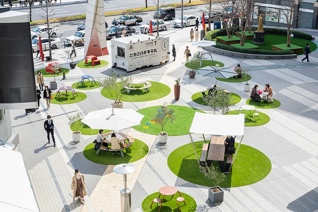 Overhead view of a public plaza in Hiroshima with bright circular patches of artificial grass, each furnished with different outdoor tables, benches, parasols, and shade structures to create spaced seating areas for visitors.