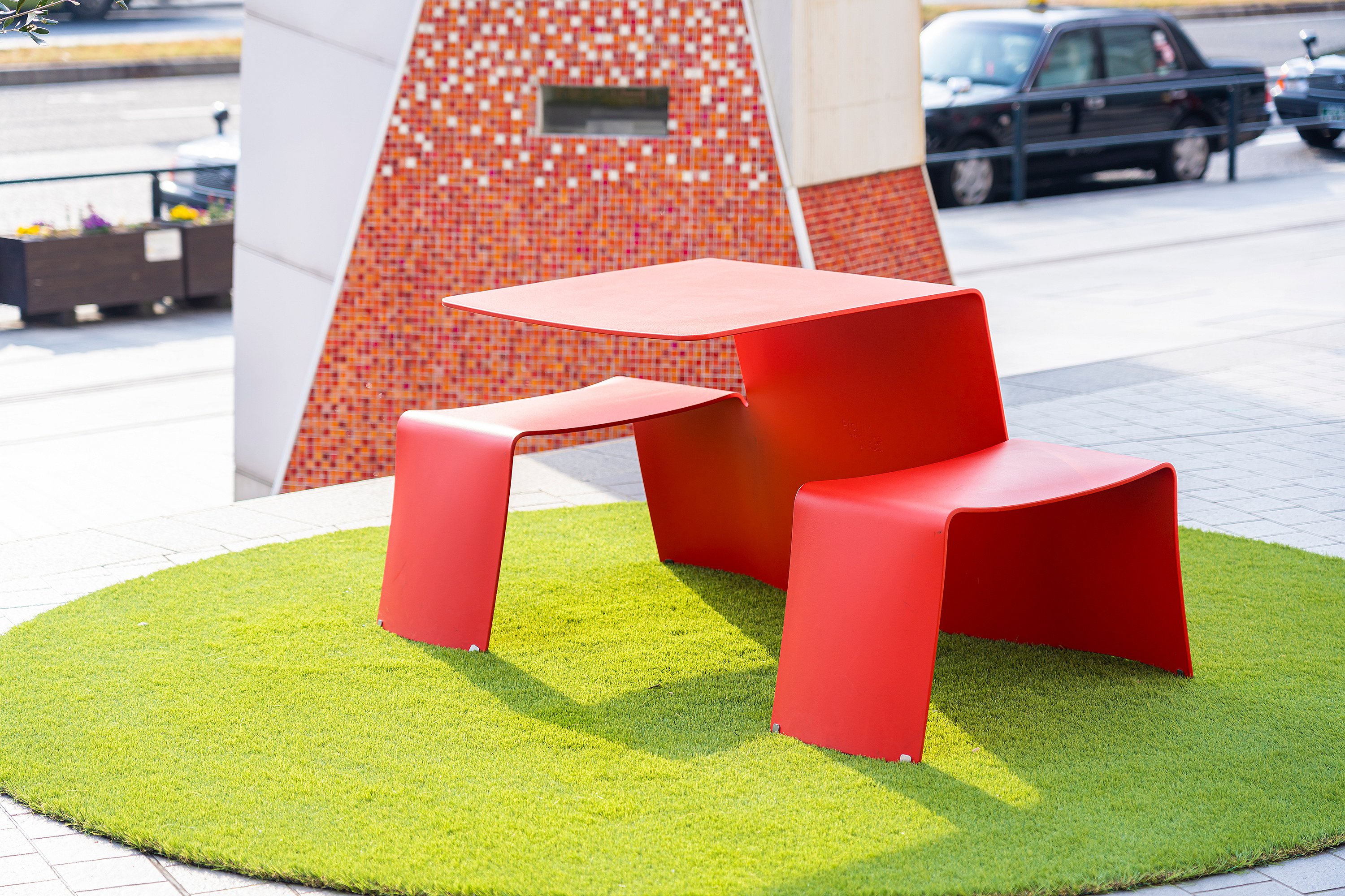 Leuchtend rote Picknickbank auf einem runden Stück Kunstrasen neben einer gefliesten Denkmalmauer auf dem Platz.