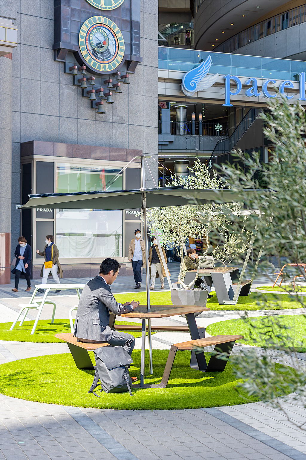 Mann im Anzug sitzt an einem runden Außentisch mit angebrachten Bänken und einem dunklen Sonnenschirm und nutzt den Tisch als ruhigen Arbeitsplatz mitten auf dem öffentlichen Platz.