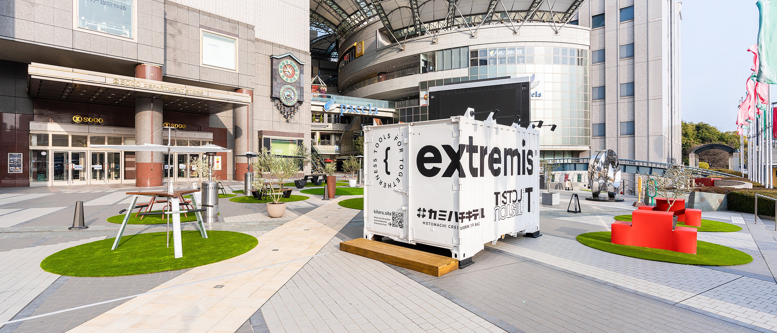 Weitblick auf die Plaza-Installation mit dem weißen Container der Marke Extremis, umgeben von separaten grünen Kreisen, die mit verschiedenen Sitzlösungen im Freien ausgestattet sind, darunter ein roter Banktisch und ein hoher Gemeinschaftstisch.