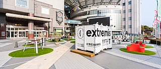 Weitblick auf die Plaza-Installation mit dem weißen Container der Marke Extremis, umgeben von separaten grünen Kreisen, die mit verschiedenen Sitzlösungen im Freien ausgestattet sind, darunter ein roter Banktisch und ein hoher Gemeinschaftstisch.