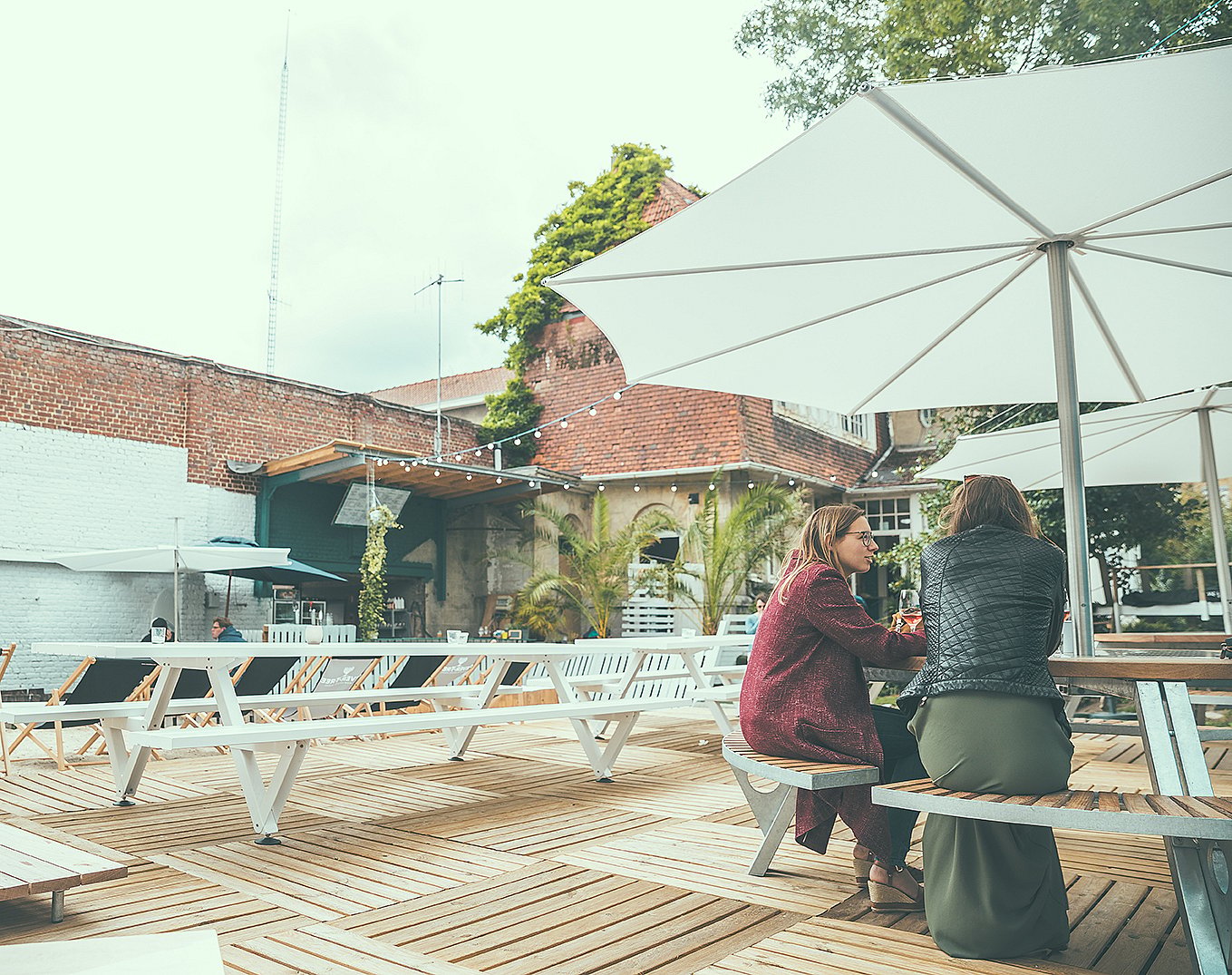 Zwei Frauen sitzen an einer Gargantua auf einer Holzterrasse, beschattet von einem großen weißen Inumbra im Innenhof der Sommerbar.