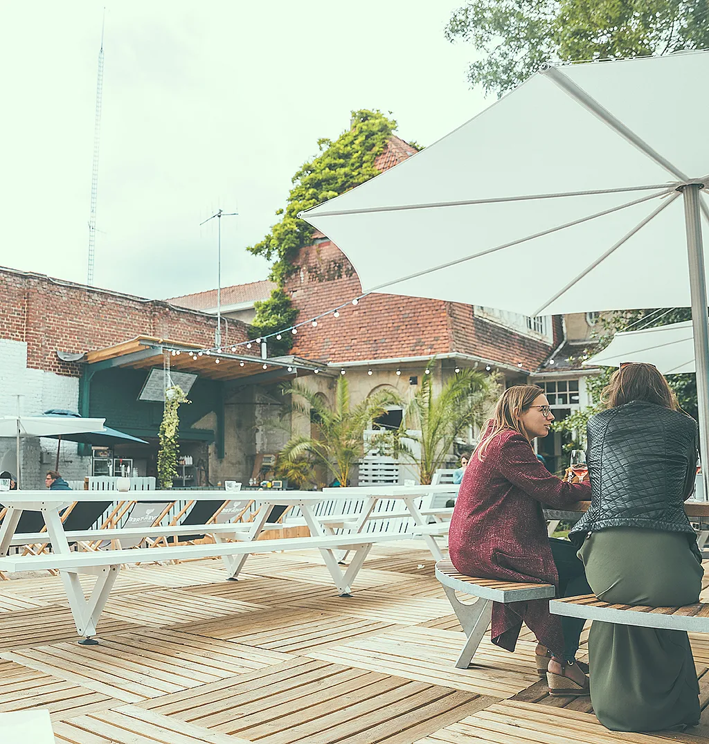 Zwei Frauen sitzen an einer Gargantua auf einer Holzterrasse, beschattet von einem großen weißen Inumbra im Innenhof der Sommerbar.