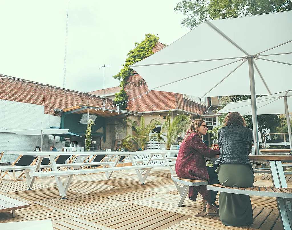 Two women seated at a gargantua on a wooden terrace, shaded by a large white inumbra in the courtyard of the summer bar.