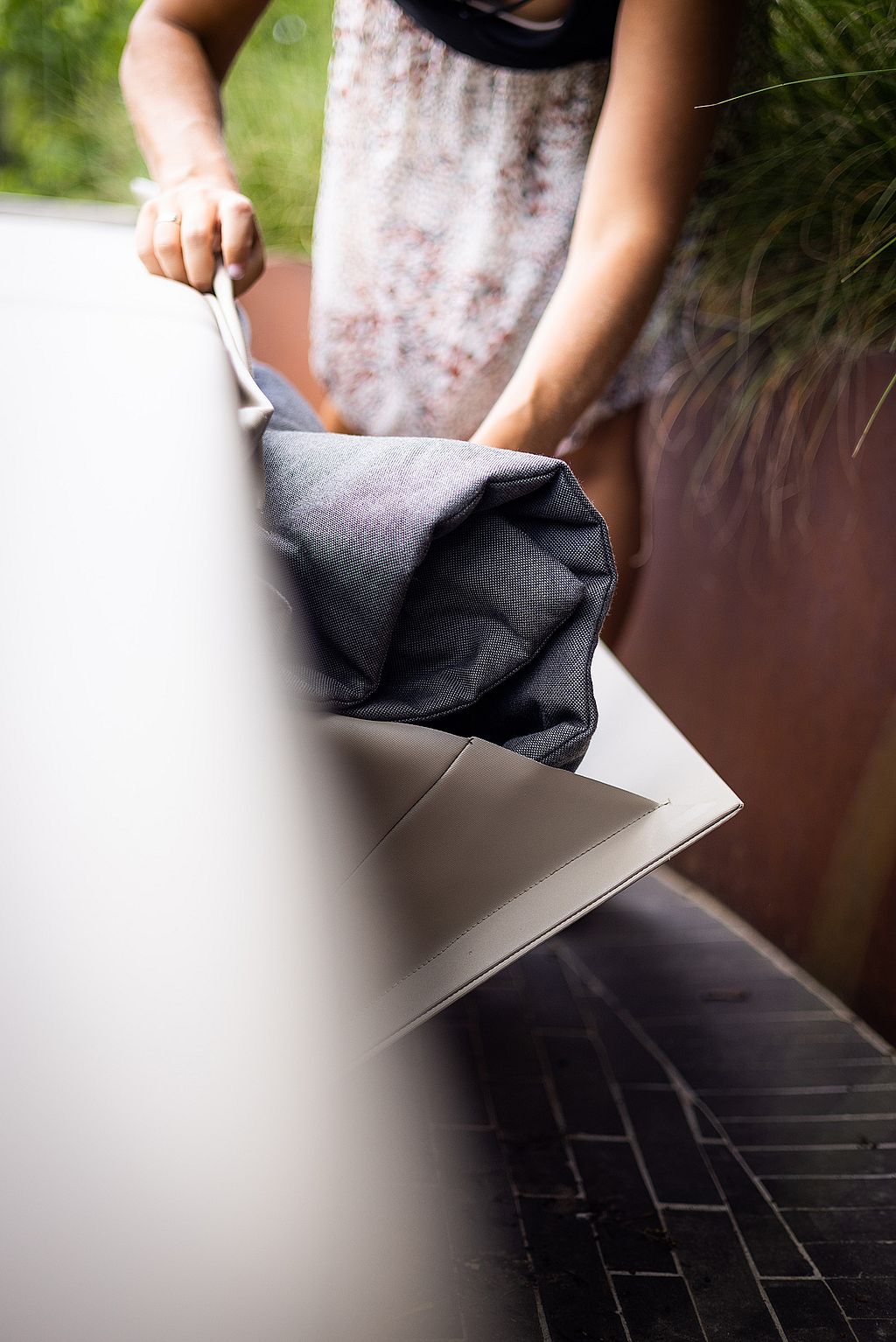 Close-up of a person lifting open the side flap of the outdoor sofa to store a folded grey blanket or cushion inside the built-in compartment.