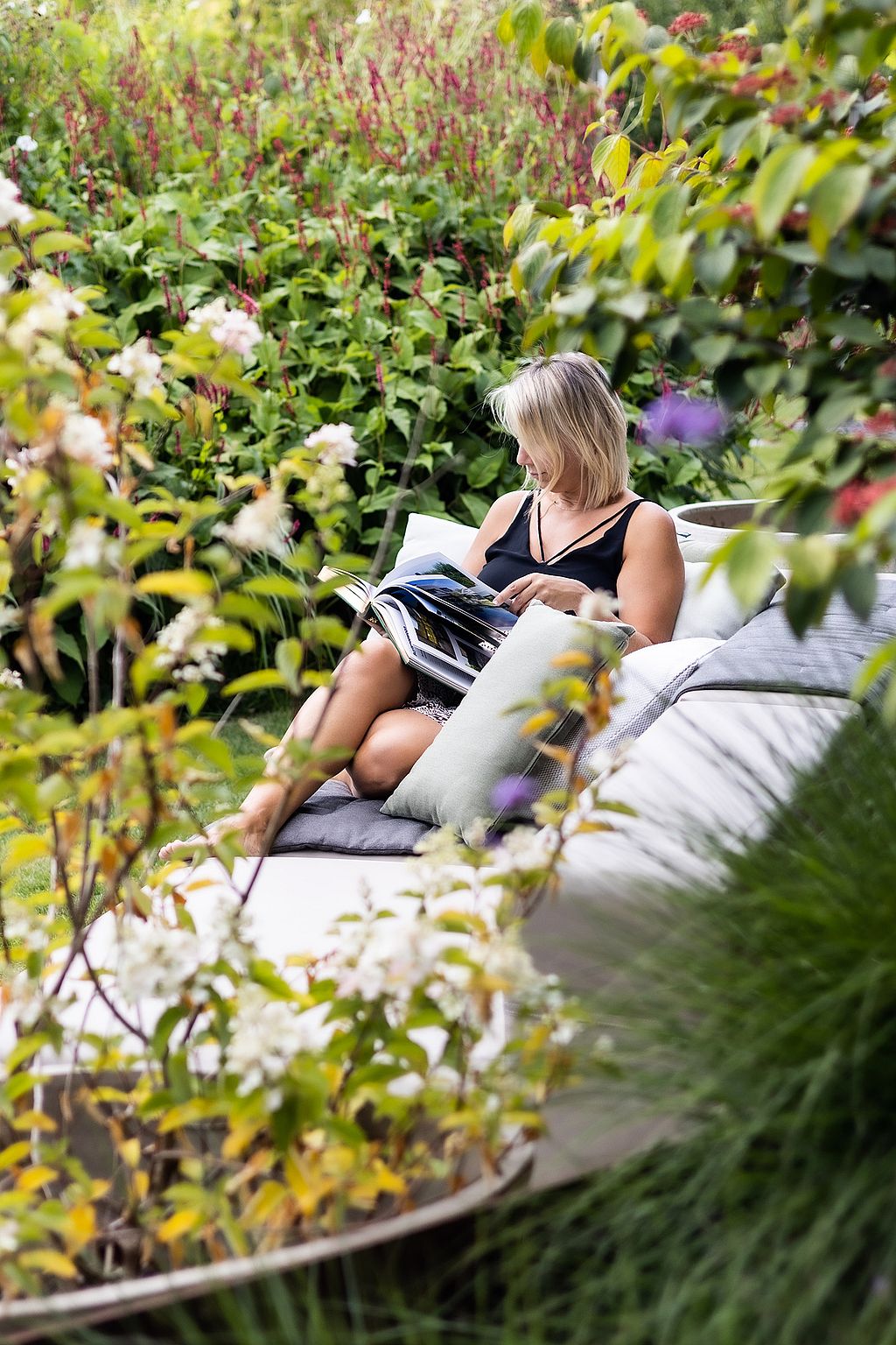 Woman reading on a modular outdoor sofa nestled in a richly planted garden, partially hidden by flowering shrubs and leafy branches in the foreground.