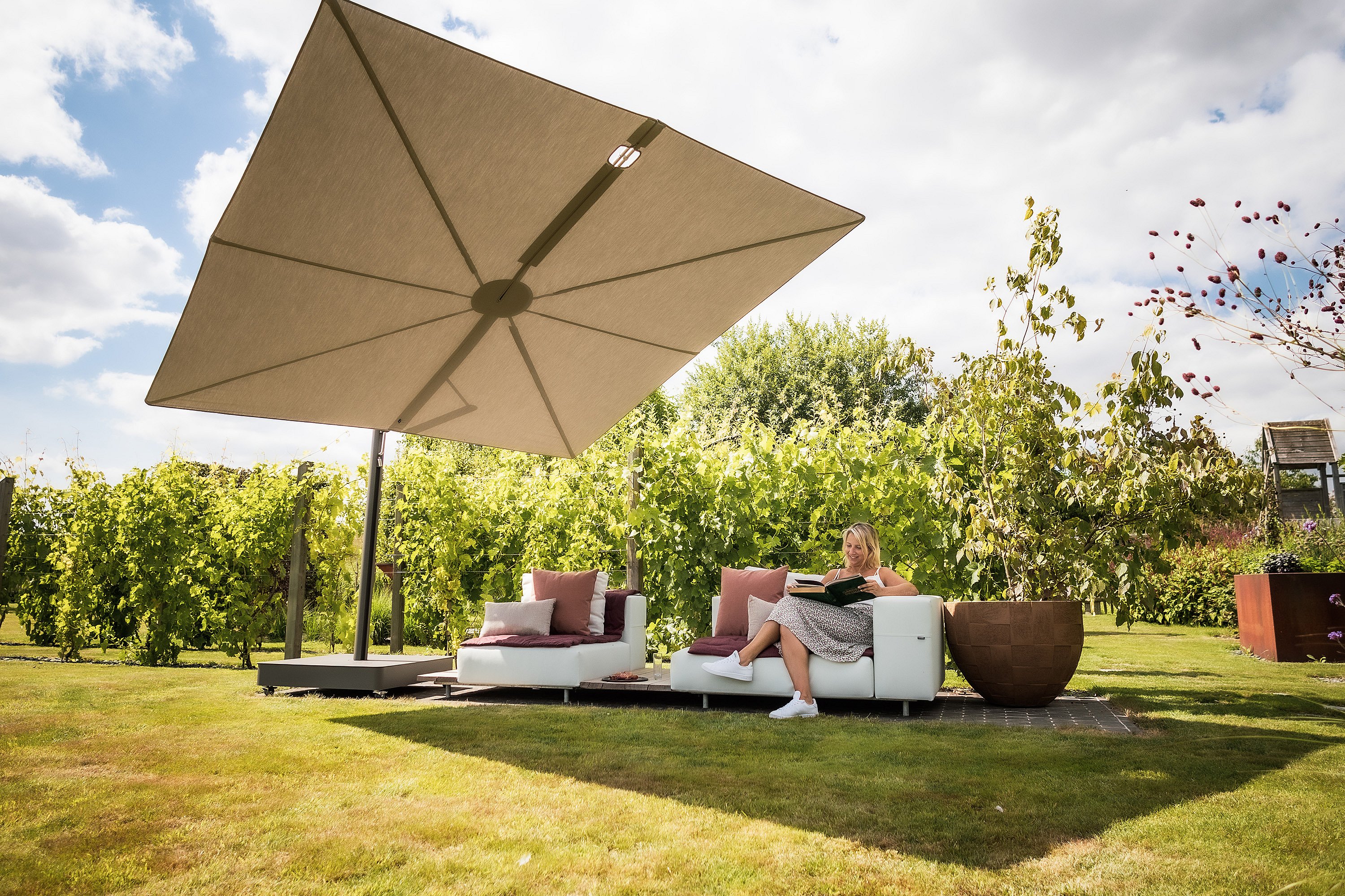 Taupe Sensu shade above a modular Walrus outdoor sofa on a lawn beside vineyard rows, where a woman sits reading among soft cushions and large corten planters.