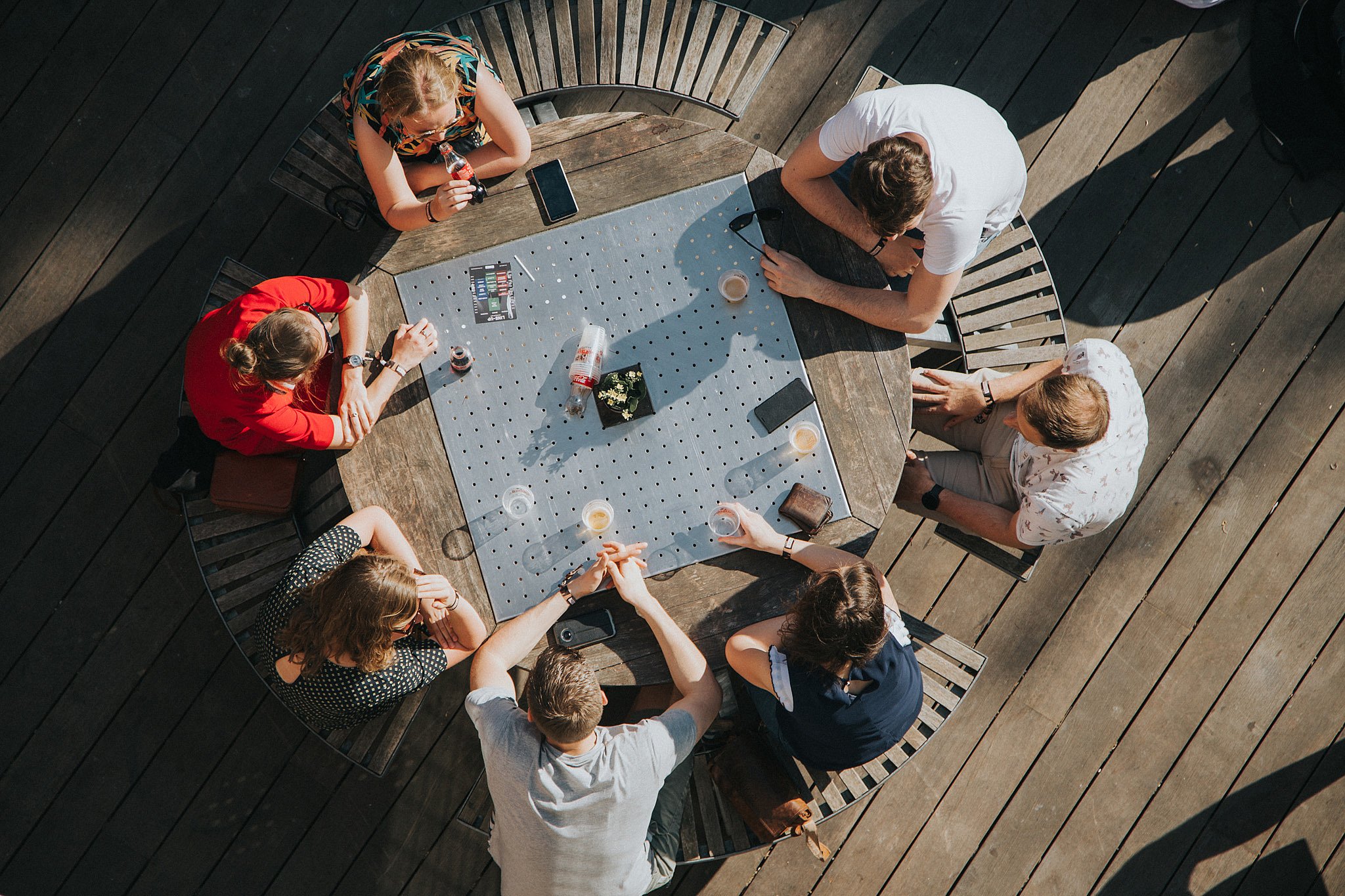 Groothoekig uitzicht op de tuin van de jeugdclub met verschillende groepen mensen verzameld rond buitentafels en banken op gras en houten terrassen tijdens een sociaal evenement.