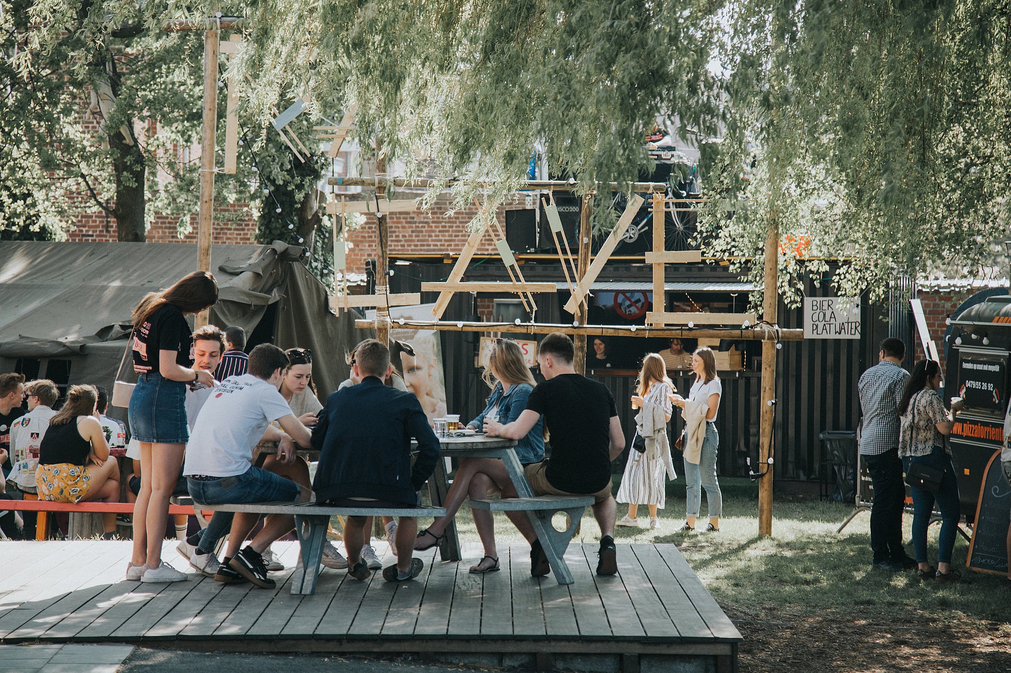 Les jeunes se sont rassemblés autour d’une table de pique-nique ronde Gargantua sur une terrasse en bois surélevée dans le jardin du club de jeunesse Den Couter, avec une petite scène extérieure et des arbres en arrière-plan.