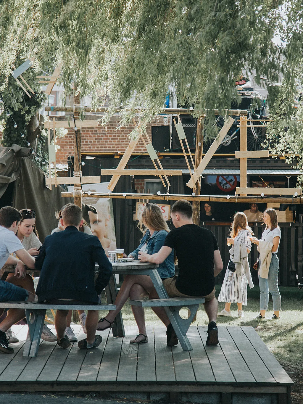 Jongeren verzamelden zich rond een ronde Gargantua-picknicktafel op een verhoogd houten terras in de tuin van jeugdclub Den Couter, met een klein buitenpodium en bomen op de achtergrond.