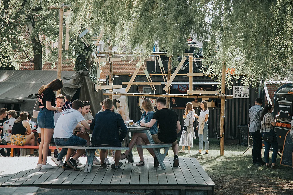 Les jeunes se sont rassemblés autour d’une table de pique-nique ronde Gargantua sur une terrasse en bois surélevée dans le jardin du club de jeunesse Den Couter, avec une petite scène extérieure et des arbres en arrière-plan.