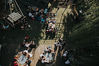 Bovenaanzicht van zes jongvolwassenen die rond een ronde Gargantua-tafel zitten met gebogen houten banken op een houten terras, met drankjes, telefoons en kleine voorwerpen verspreid over het tafelblad.
