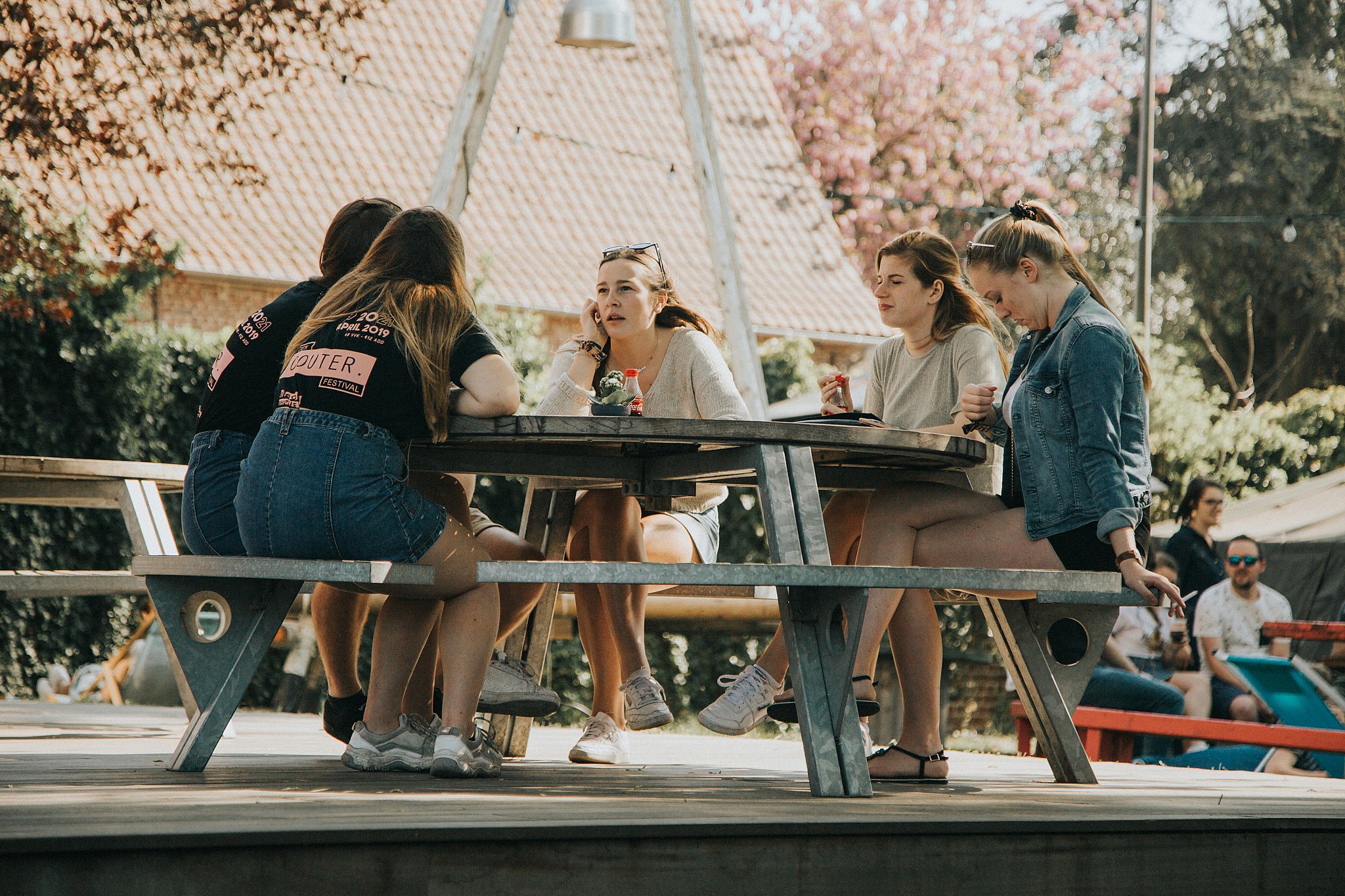 Vijf jonge vrouwen zitten samen aan een ronde Gargantua-picknicktafel op een houten platform, kletsen in de zon met bloeiende bomen en de tuin van de jeugdclub erachter.