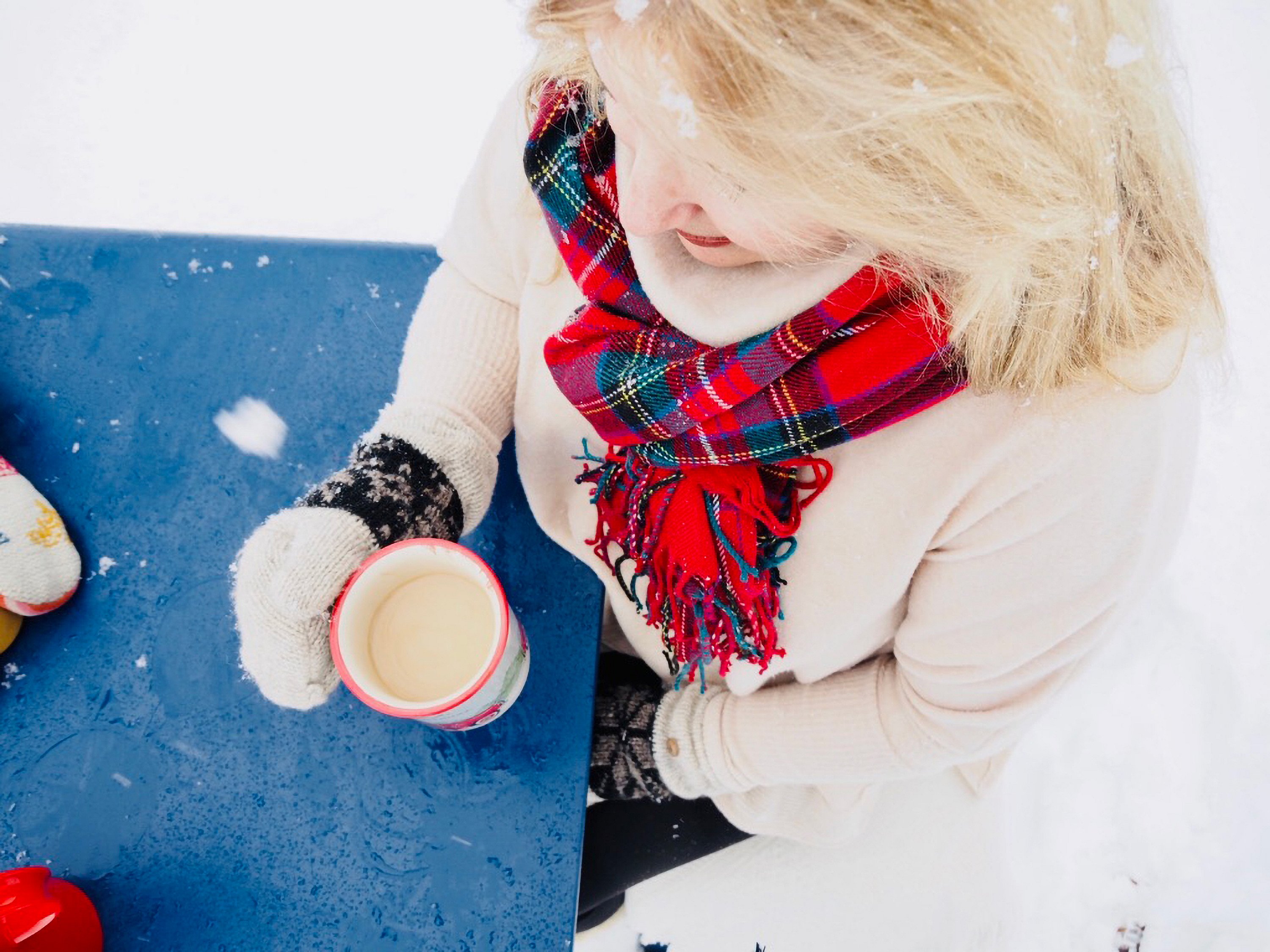 Close-up from above of a woman in mittens and a red tartan scarf holding a mug beside a wet blue tabletop dusted with snow.