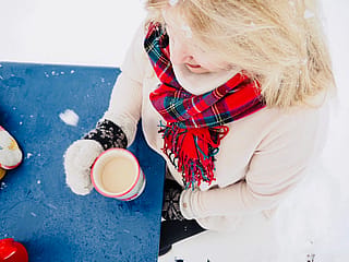 Close-up from above of a woman in mittens and a red tartan scarf holding a mug beside a wet blue tabletop dusted with snow.