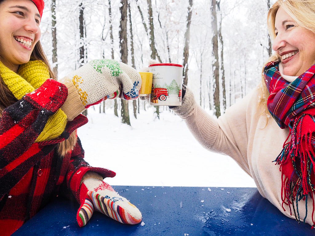Close-up of two women toasting hot drinks across a blue outdoor table, with colorful knitted mittens and a snowy woodland in the background.