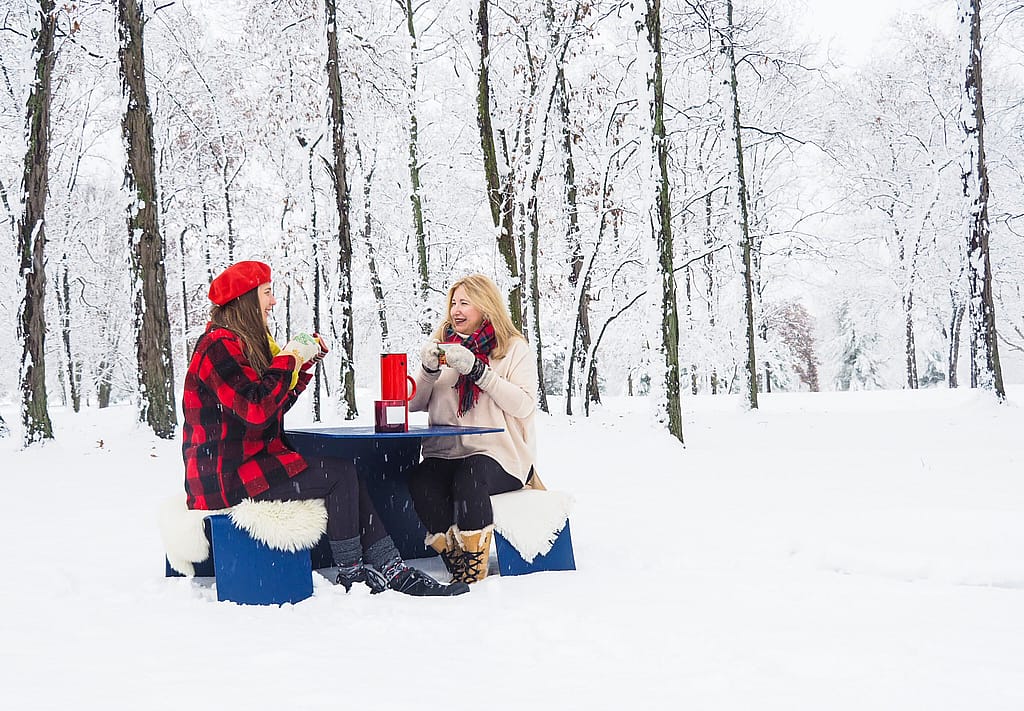 Two women bundled in scarves, mittens, and boots sit at a blue outdoor table in deep snow, smiling over hot drinks with bare winter trees behind them.