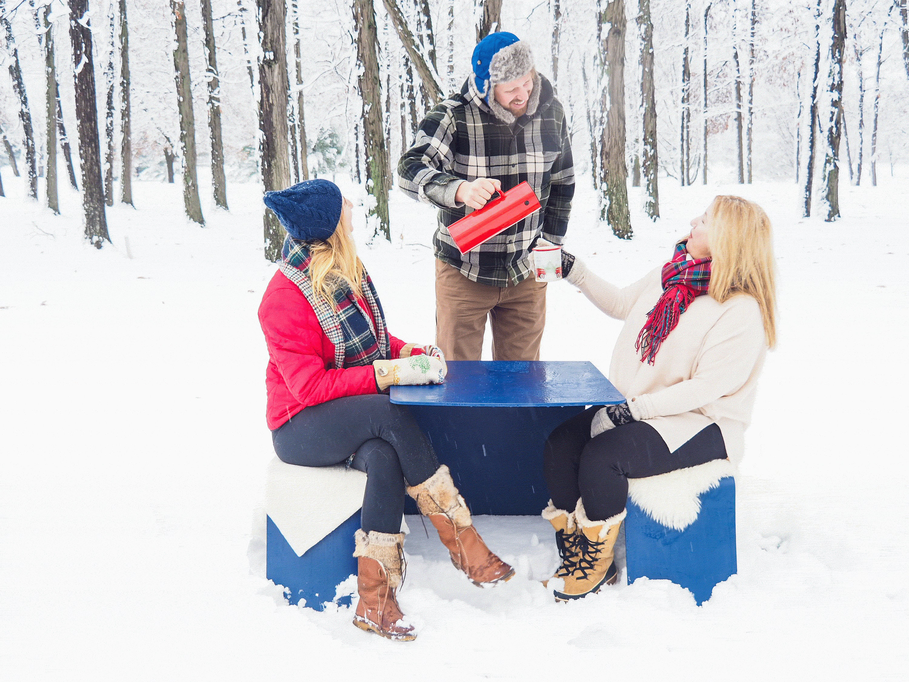 Trois personnes en vêtements d’hiver se rassemblent autour d’une table de pique-nique extérieure bleue dans une forêt enneigée, versant et tenant des boissons chaudes tout en étant assises sur des bancs drapés de peaux de mouton blanches.