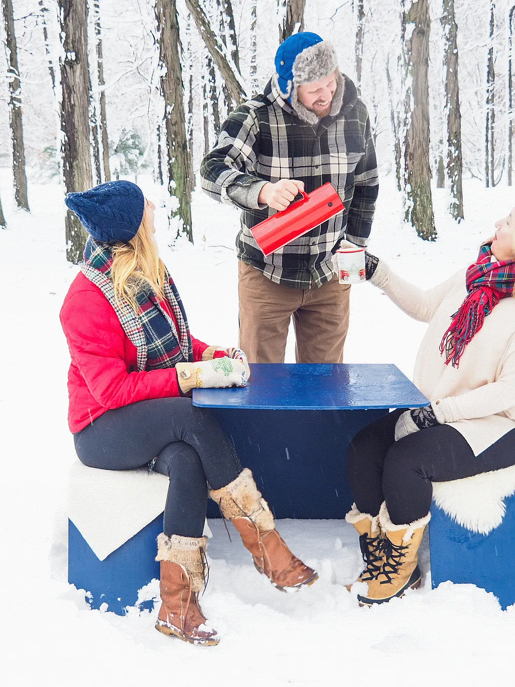 Trois personnes en vêtements d’hiver se rassemblent autour d’une table de pique-nique extérieure bleue dans une forêt enneigée, versant et tenant des boissons chaudes tout en étant assises sur des bancs drapés de peaux de mouton blanches.