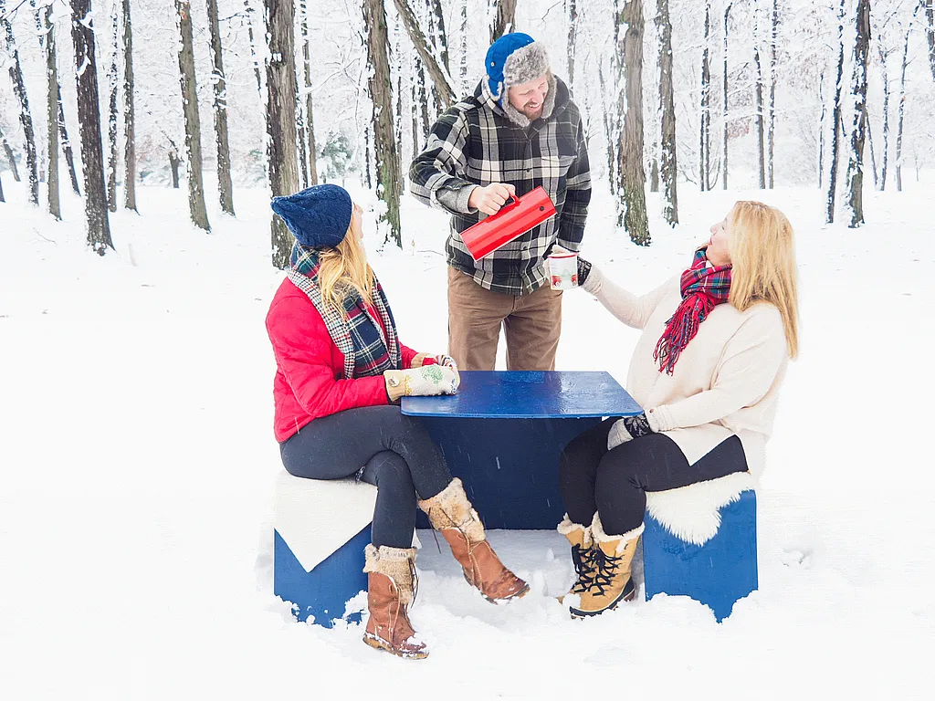 Three people in winter clothing gather around a blue outdoor picnic table in a snow-covered forest, pouring and holding hot drinks while sitting on benches draped with white sheepskins.