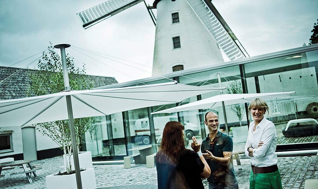 Drie mensen kletsen op de geplaveide binnenplaats van het d-hotel onder grote witte parasols, met de historische witte windmolen en een glazen hoteluitbreiding erachter.