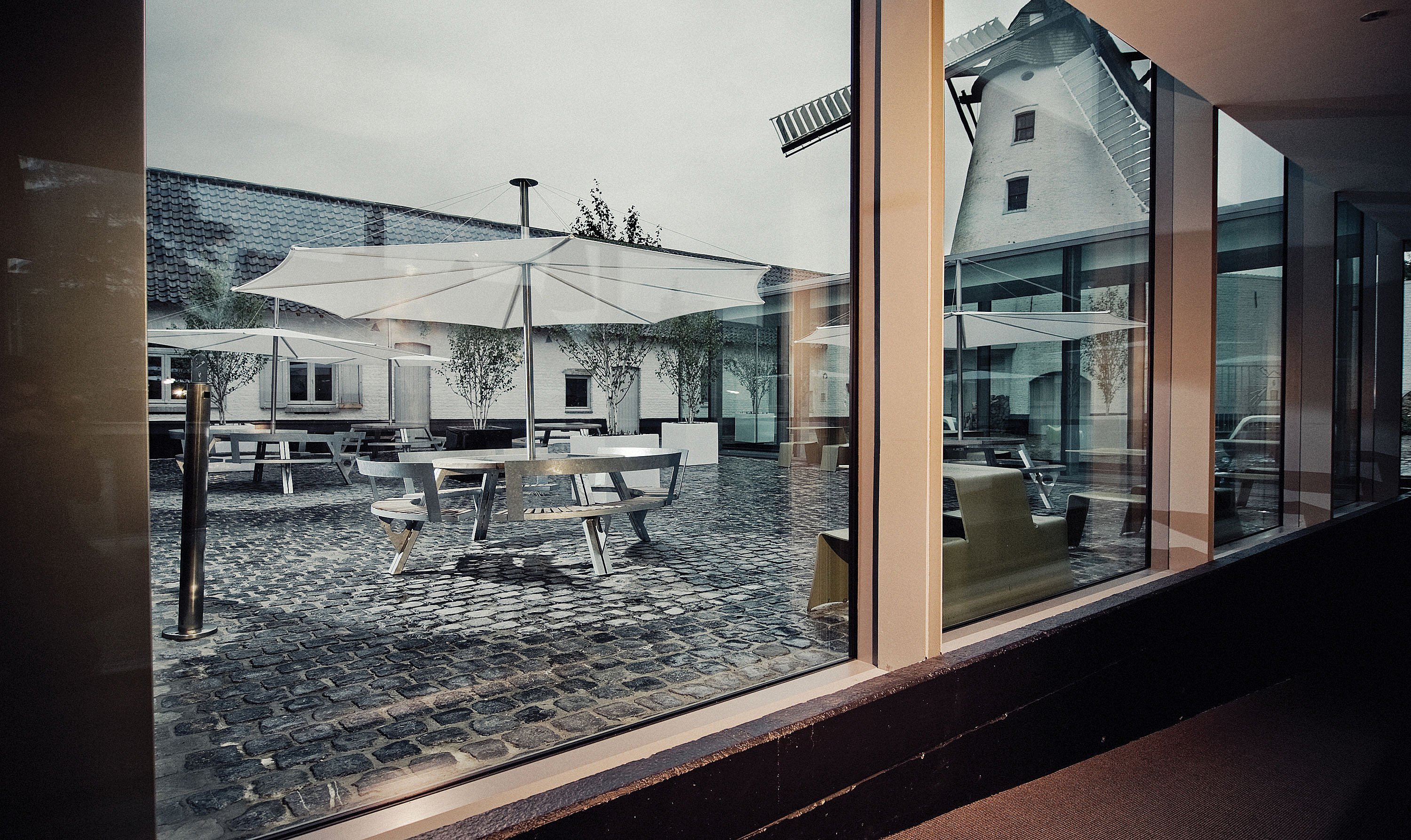 Binnenaanzicht door een groot hotelraam op de geplaveide binnenplaats, met ronde buitentafels en witte parasols omlijst door de moderne glazen gevel en de oude windmolen daarachter.
