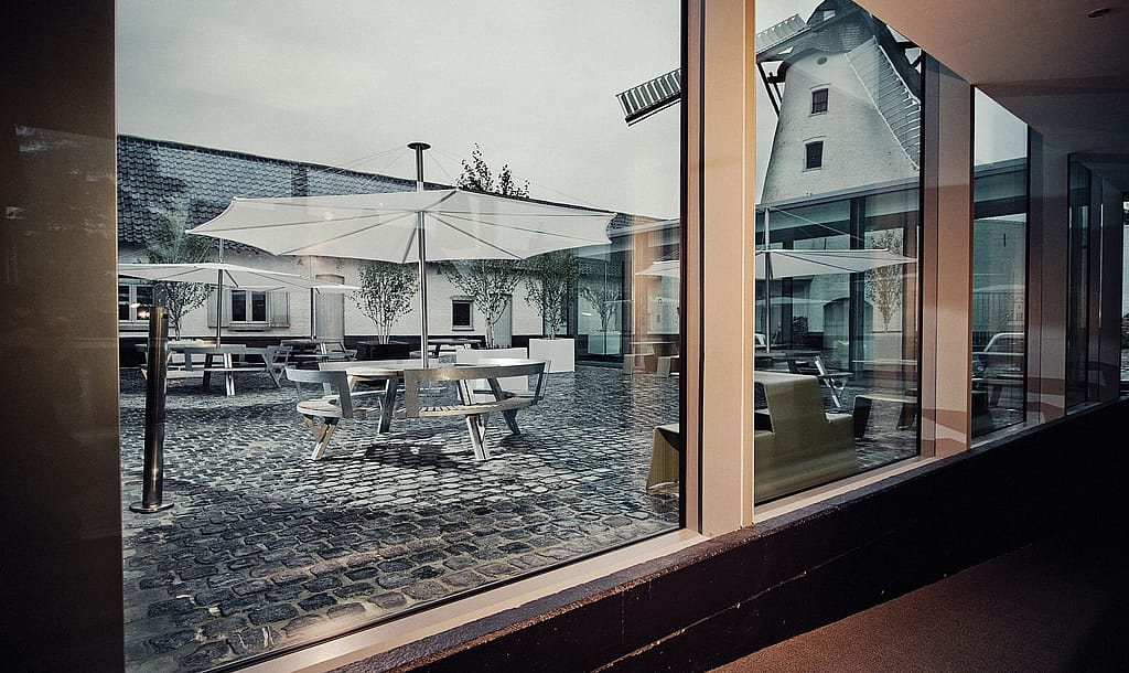 Binnenaanzicht door een groot hotelraam op de geplaveide binnenplaats, met ronde buitentafels en witte parasols omlijst door de moderne glazen gevel en de oude windmolen daarachter.