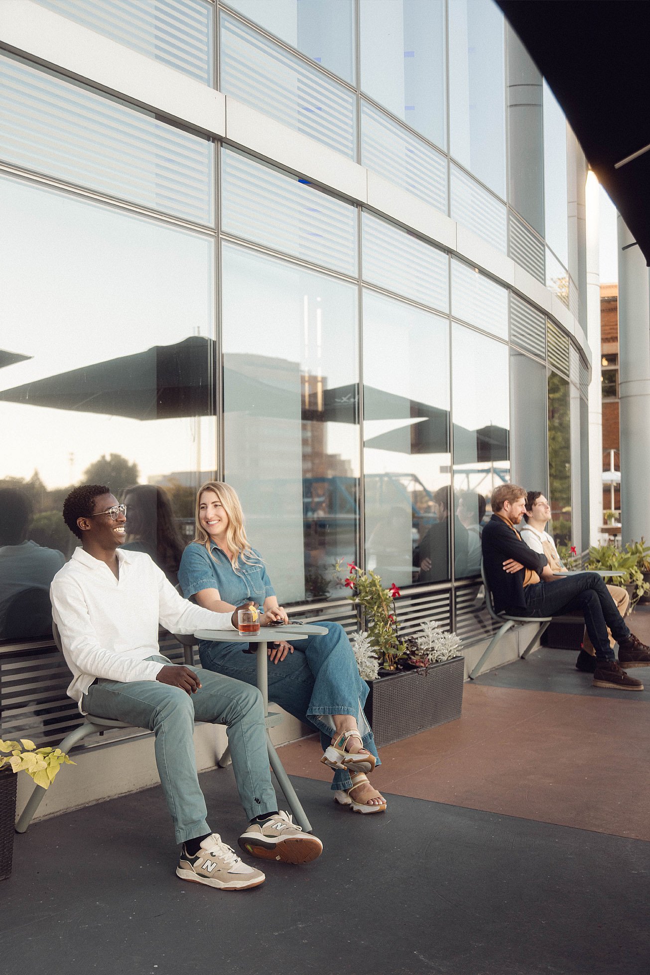 Deux clients de l’hôtel sont assis à une petite table ronde Bistroo à côté de la façade en verre réfléchissant, tandis que deux autres couples se détendent plus loin le long du banc de la terrasse.