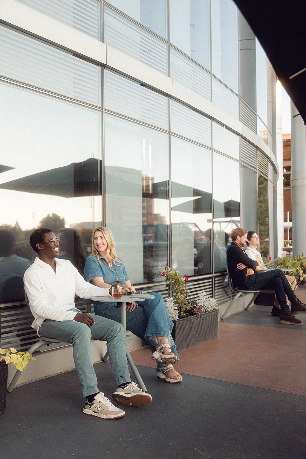 Deux clients de l’hôtel sont assis à une petite table ronde Bistroo à côté de la façade en verre réfléchissant, tandis que deux autres couples se détendent plus loin le long du banc de la terrasse.