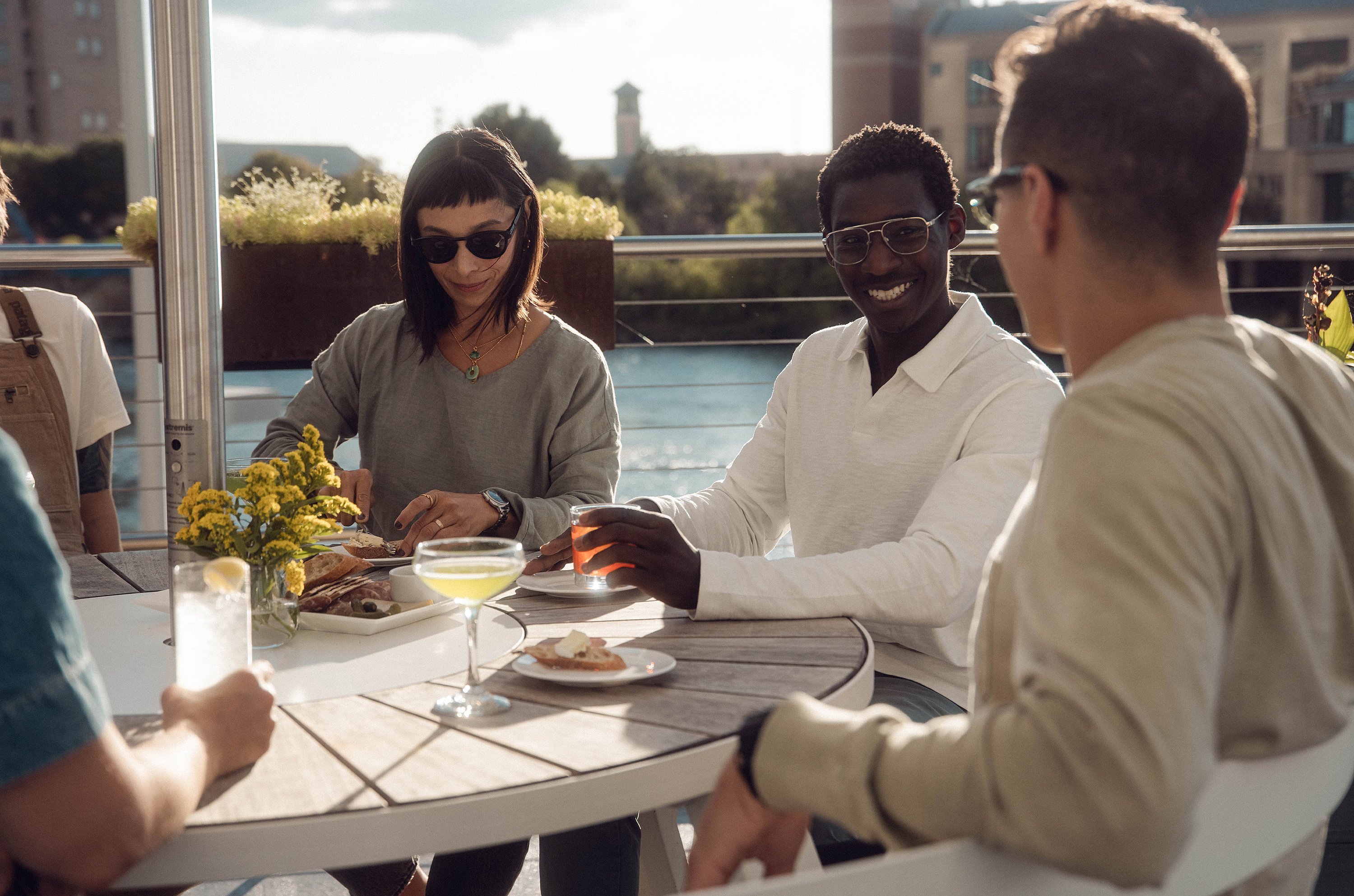 Gros plan de quatre invités assis autour d’une table ronde blanche en plein air avec des cocktails, des assiettes et des fleurs, profitant du coucher de soleil sur la terrasse sur le toit.