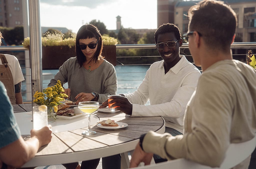 Gros plan de quatre invités assis autour d’une table ronde blanche en plein air avec des cocktails, des assiettes et des fleurs, profitant du coucher de soleil sur la terrasse sur le toit.